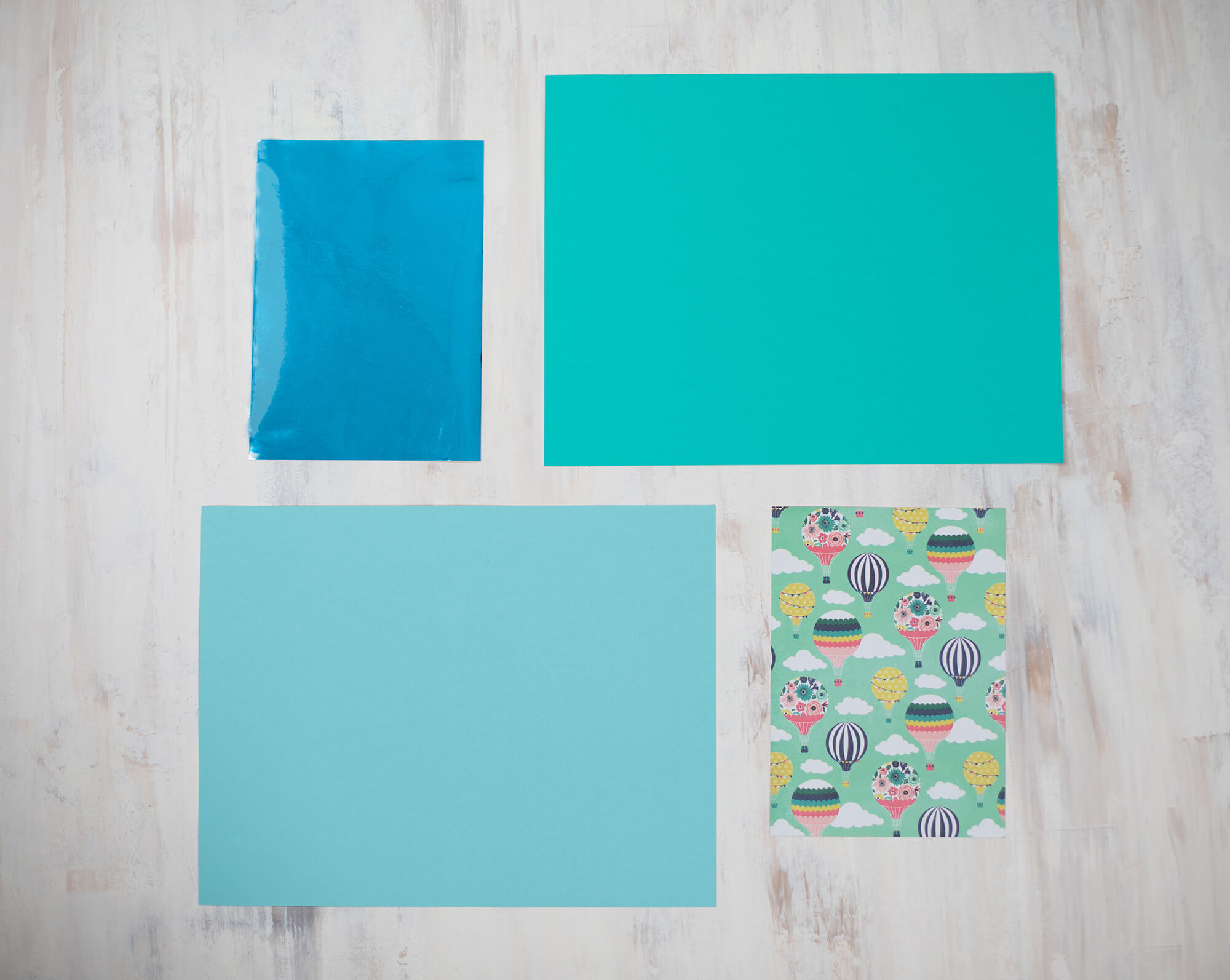Four pieces of paper arranged on a light wooden surface: one bright blue glossy sheet, one large turquoise sheet, one smaller light blue sheet, and one patterned sheet with colorful hot air balloons and clouds on a mint green background.