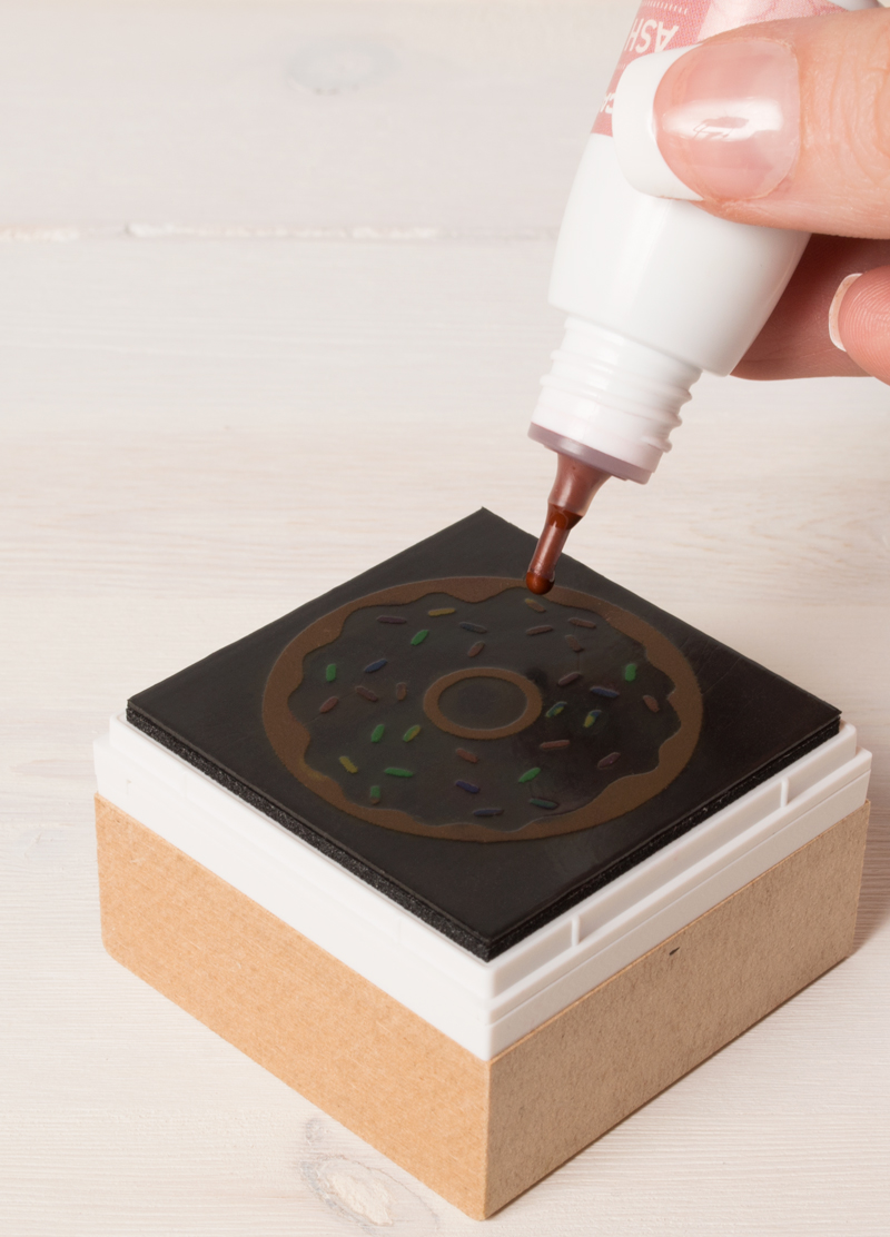 A person applying brown ink to a stamp with a donut design, which is mounted on a wooden and plastic stamp block, placed on a light wooden surface.