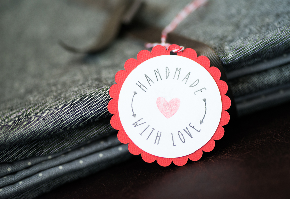 A close-up of a red scalloped-edged tag with the text 'Handmade with Love' and a heart symbol, attached to a stack of gray fabric.