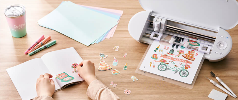 A person using a cutting machine to create various colorful paper cutouts on a wooden desk. The desk is also adorned with a pastel travel mug, a stack of pastel-colored papers, markers, and craft tools. The person is arranging cutouts in a scrapbook, with