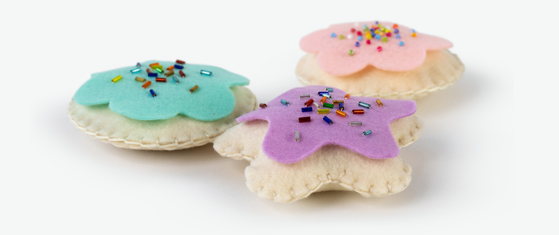 Three handmade felt cookies with pastel-colored frosting and colorful bead sprinkles, arranged on a white surface.