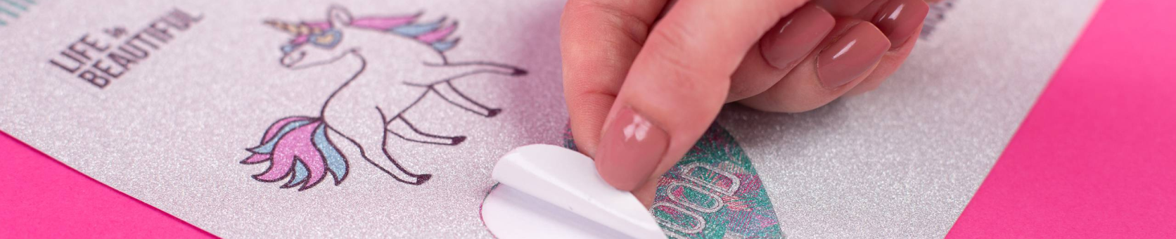 Close-up of a person's hand peeling a glittery sticker from a sheet, with a unicorn illustration and the text 'Life is Beautiful' on a pink background.