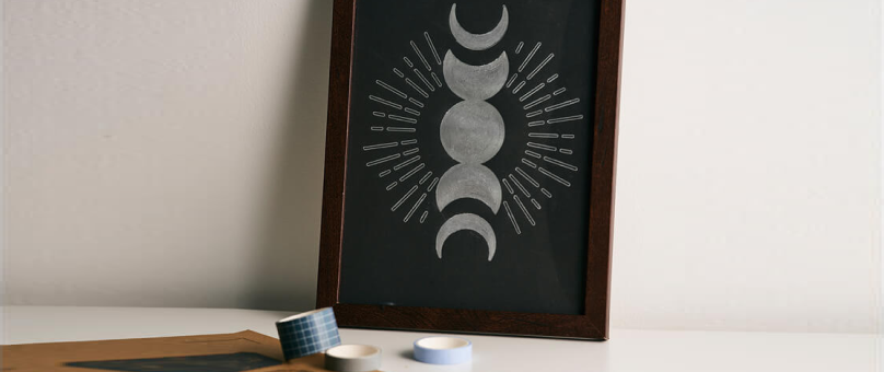 A framed artwork depicting the phases of the moon, with a series of crescent and full moons arranged vertically and surrounded by radiating lines, placed on a white surface. In front of the frame are three rolls of decorative tape in different patterns an