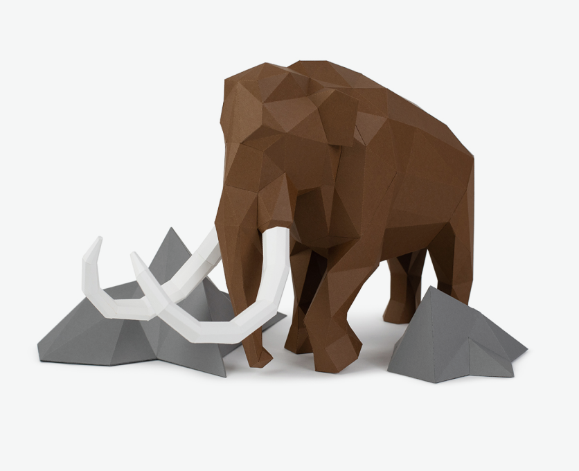 3D paper model of a woolly mammoth with white tusks, standing on a white surface with grey geometric rocks around it.