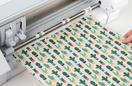 Image of a Cameo cutting machine with a cutting mat loaded with pink paper shapes, next to a vase of pink and green paper flowers.