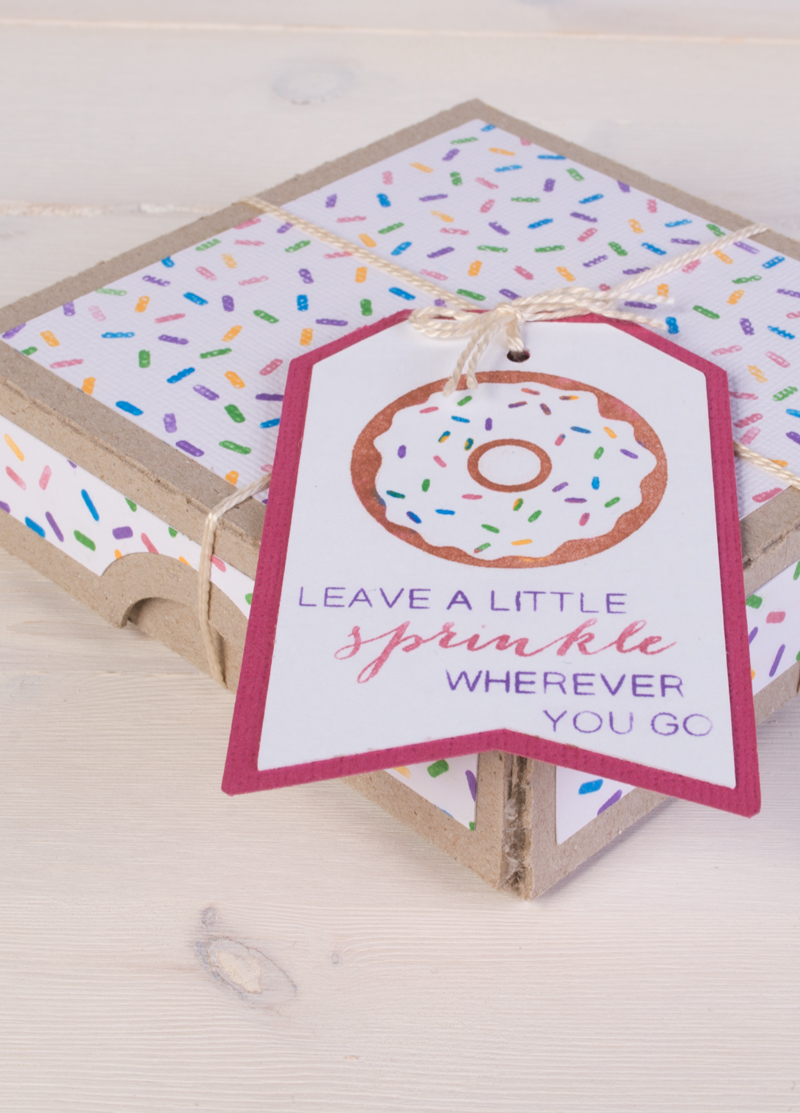 A decorated gift box wrapped with twine and a tag featuring a donut illustration and the phrase 'Leave a little sprinkle wherever you go.' The box is adorned with colorful sprinkle patterns, placed on a light wooden surface.